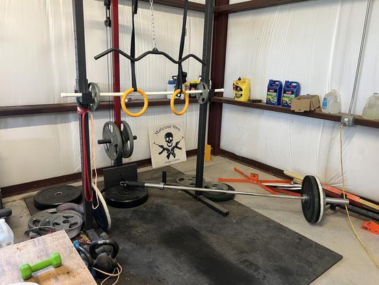 Bar bell, dumbbells, and kettlebells on rubber flooring with a rack holding gymnast rings and a lat pulldown bar