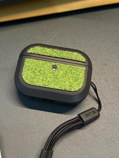 Picture of a small black dbrand air pods pro 2 case, with a black lanyard attached to it.

On the front panel there is a green fabric green that looks like fake grass.