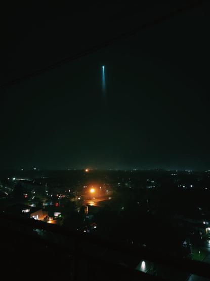 View from an apartment at night of an unidentified flying object shining a light downward hovering above the city.