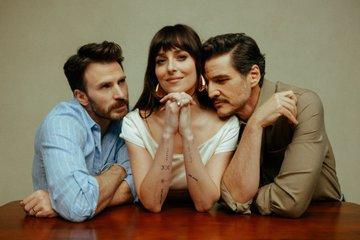 Dakota Johnson sits at a table. On her left side sits Chris Evans and admire her. On the right side sits Pedro Pascal and has his chin bedded on Dakota's shoulder.