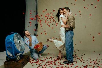 Dakota Johnson and Pedro Pascal hug each other. Chris Evans kneels next to a big fan and trows red rose petals in the air so that the fly around Pedro and Dakota.