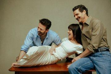 Dakota half sits/half lies on a table. Pedro is supporting her head which she has laid against his chest. Chris Evans sits next to her. All three are laughing out loud.