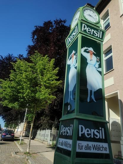 Das Bild zeigt eine grüne Persilstanduhr. Im Hintergrund ein Haus und Bäume. 