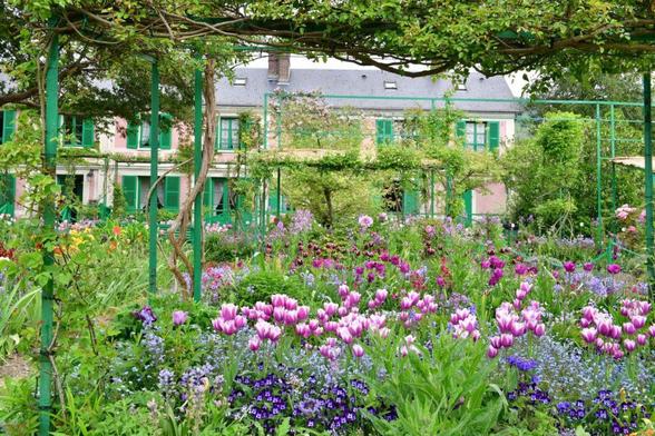 A big one with lots of green. Lilac small flowers in the foreground and behind that some bigger flowers white with some darker lilac. At the end of the garden is a big house.
