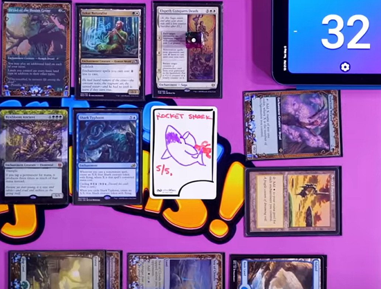 A screenshot of a EDHinks playmat with Magic cards on it. It has an InfiniTokens dry erase token in the center that says "Socket Shark 5/5" with a roaring shark with a flaming rocket strapped to its back drawn on it. 