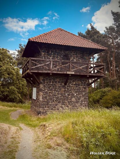 Rekonstruierter römischer Wachturm bei Grüningen mit dunklem Bruchsteinmauerwerk und Ziegeldach, aufgenommen aus leicht schräger Perspektive. Ein schmaler Pfad führt durch hohes Gras direkt auf den Turm zu. Die Szene wirkt ruhig und geschichtsträchtig unter blauem Himmel mit einzelnen Wolken.