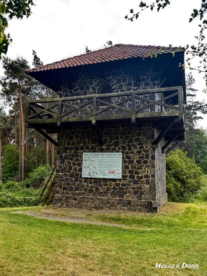 Frontalansicht des rekonstruierten Limesturms bei Grüningen, mit dunklem Naturstein und umlaufendem hölzernen Balkon. Im unteren Bereich eine weiße Infotafel mit der Aufschrift „Wachturm am Posten 49“. Der Turm steht am Rand eines Waldstücks und vermittelt den Eindruck römischer Präsenz inmitten der Natur.