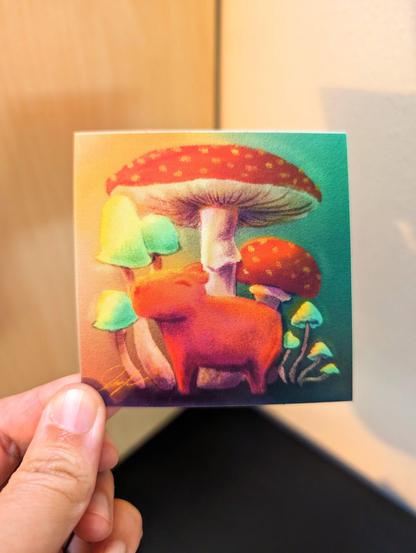 Photo of a left hand holding a square sticker. The sticker is a matte drawing of a capybara standing with mushrooms.