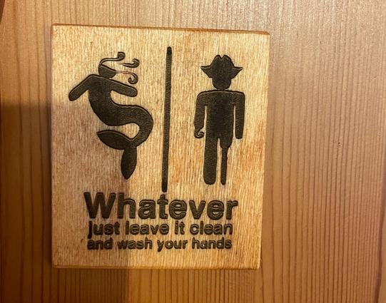 A sign on a wooden door, laser-engraved on plywood. It shows the silhouette of a mermaid on the left and the silhouette of a pirate with a hook for one hand and a wooden leg, between them a vertical line. The word „Whatever“ is written below in big letters, below that the words „just leave it clean and wash your hands“.