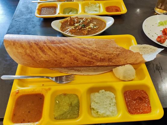 A dosa (similar to a large thin pancake, crisp on one sie) on a large yellow rectangular plastic canteen tray the tray has an upper section with the dosa, four lower sections with different sauces. The dosa is wrapped into a tube.