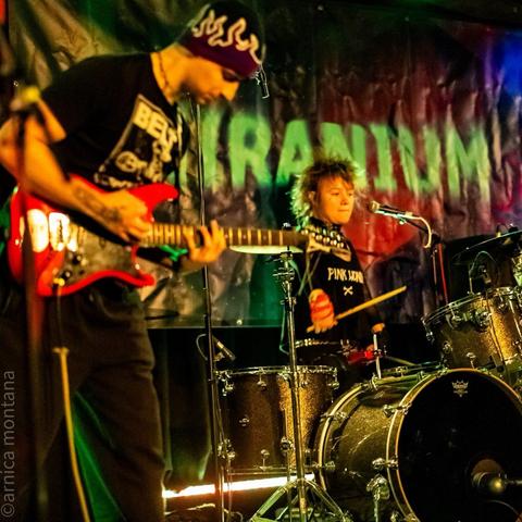 A band performs on stage, featuring a guitar player in a black t-shirt and a beanie, and a drummer with tousled hair wearing a black "Pink Wonder" shirt. The background includes a banner with the word "NIRANIUM"