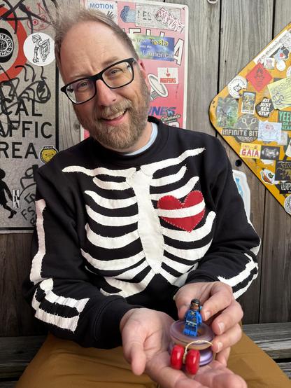 A man in a bar posing for the camera while holding a yo-yo and a customized Lego mifigurenifigure in his hand. The man is Doc Pop, and he is wearing a black sweatshirt with a skeleton design on it that was handstitched using applique techniques and white embroidery thread.