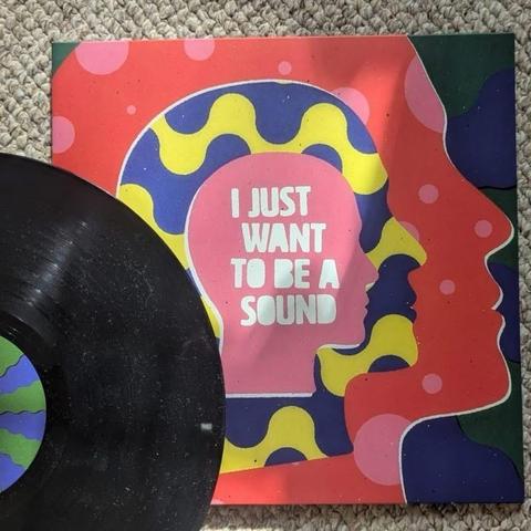 Eine Schallplatte ist teilweise neben ihrem farbenfrohen Albumcover zu sehen. Das Cover zeigt abstrakte Formen und leuchtende Farben, wobei der Satz "I JUST WANT TO BE A SOUND" in weißer Schrift vor einem rosafarbenen Hintergrund zu lesen ist.