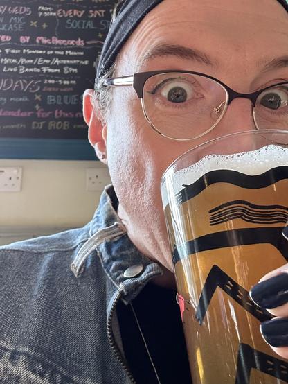 A selfie of me at the pub. I’m drinking a pint of beer. The nails on my hand holding the beer are painted deep blue. I’m wearing glasses, behind which I’m wearing subtle black eyeliner with a small flick. My hair is pulled back in a black hairband, and I’m wearing a denim jacket. The photo is close enough that part of my face is out of shot. Behind be is a blackboard with various events advertised.