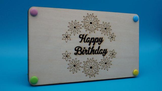 Rechteckige Holzkarte mit graviertem „Happy Birthday“-Text in einem floralen Kranz, farbig verschlossen mit bunten Steckpunkten.