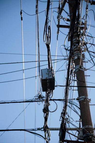 A tangle of black and silver power lines stretches against a clear blue sky, with a gray utility box prominently positioned among the wires.