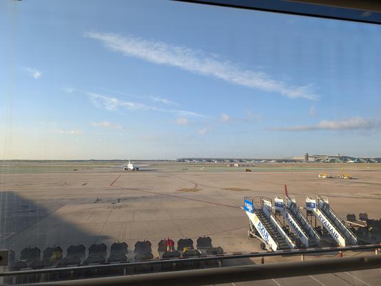 An Airport rolling field. In the back a plane Rolls. In Front you can See a bunch of mobile stairs an luggage cars