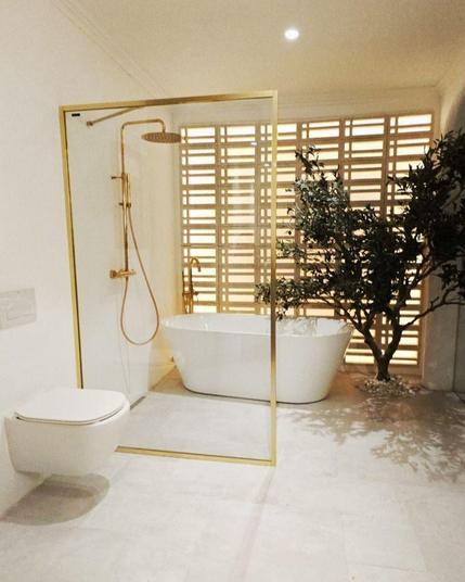 Baño moderno en casa Decor 2025 con bañera independiente de porcelana blanca, mampara de cristal con perfilería y grifería en dorado e inodoro suspendido