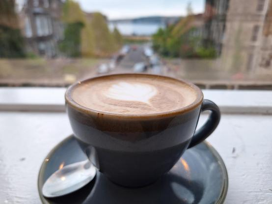 Coffee cup with flower latte art on a window sill looking out on a road leading away towards some water or so (actually the River Tay)