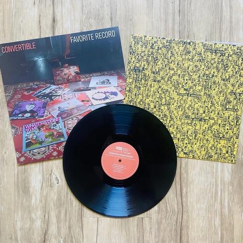 Ein Albumcover für "Convertible - Favorite Record" wird neben einer Vinyl-Schallplatte gezeigt. Das Cover zeigt ein Foto von verschiedenen Schallplatten auf einer gemusterten Oberfläche. Die Schallplatte hat ein rotes Etikett. Die Rückseite des Covers ist gelb mit schwarzem Text.
