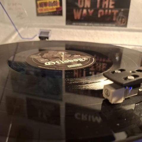 Nahaufnahme einer sich drehenden schwarzen Schallplatte auf einem Plattenteller mit einem zentralen Etikett mit verschlungenen Mustern. Im Hintergrund sind teilweise sichtbare Poster oder Kunstwerke an der Wand zu sehen.