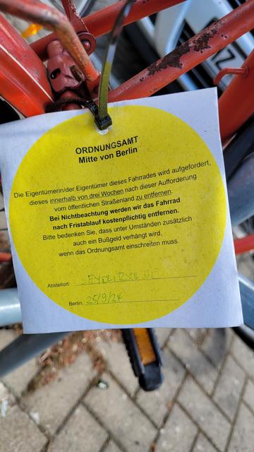 Zettel, mit Kabelbinder an einem Schrottrad befestigt, das einen öffentlichen Fahrradbügel blockiert: "ORDNUNGSAMT
Mitte von Berlin
Die Eigentümerin/der Eigentümer dieses Fahrades wird aufgefordert, dieses innerhalb von drei Wochen nach dieser Aufforderung vom öffentlichen Straßenland zu entfernen.  Bei Nichtbeachtung werden wir das Fahrrad nach Fristablauf kostenpflichtig entfernen. 
Bitte bedenken Sie, dass unter Umständen zusätzlich
auch ein Bußgeld verhängt wird, 
wenn das Ordnungsamt einschreiten muss. 
Abstellort:
Seydlitzstr., Berlin, 25/9/24". 