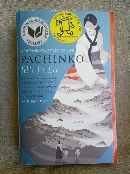 Book: Pachinko by Min Jin Lee. Light blue cover background, silhouette of a woman in kimono, the kimono makes yet another cover picture - a landscape with hills and valleys and a red sun and a woman and two children in the foreground. 