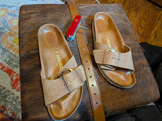 A pair of Birkenstock sandals and a brown belt on a brown ottoman with the Victorinox Swiss Champ Swiss Army Knife's Awl tool deployed. 