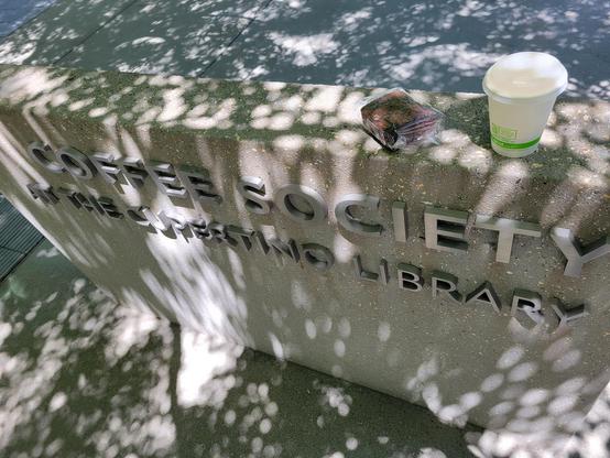 Photo of mid-day outdoor patio, showing shaded low concrete wall with silver block letters reading "Coffee Society" (large font) and "At the Cupertino Library" (small font). The light is dappled by deciduous trees. On top of the wall at right is a brownie wrapped in plastic and a to-go coffee cup.