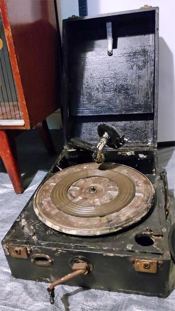 An old wind-up gramophone record player. It is portable with a lid and handle The player is rusty and has been repainted. 