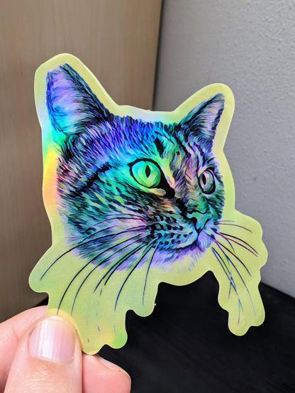 Photo of a left hand holding a diecut sticker. The sticker is a holographic tabby cat drawing.