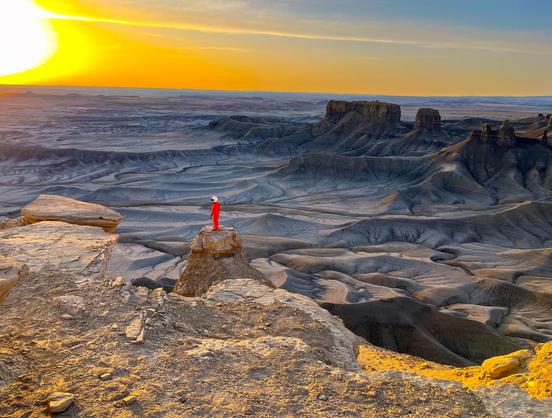 A lone person in a red suit stands on a rock formation overlooking a surreal, eroded desert landscape at sunrise. The sky glows golden on the left while the terrain below shows intricate textures and patterns in shades of gray and beige.