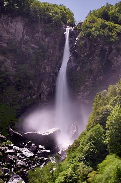 A long-exposure shot of Cascata di Foroglio, capturing the waterfall’s fierce, roaring energy in a form that looks ethereal and gentle, like a bride’s veil shimmering in the sunlight. The water softly blankets the stones below, framed by fresh, sunlit greenery, creating a peaceful contrast to the waterfall’s true, resounding force.