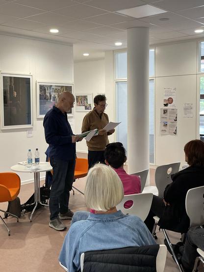 Zwei Männer stehen, lesend vor Publikum im Hintergrund Bilderrahmen mit Fotos der Ausstellung.