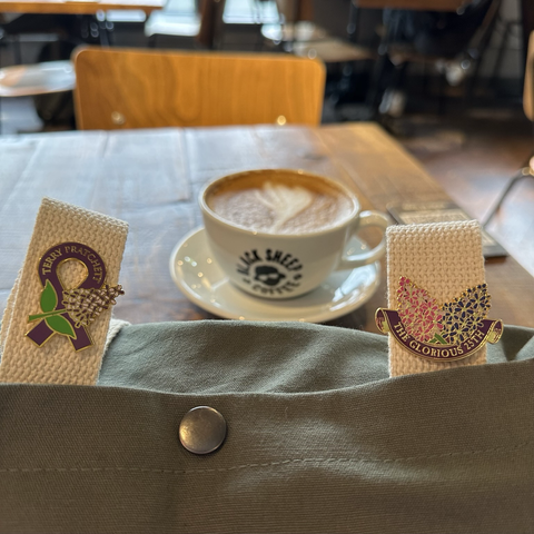 My bag, with the woven straps - pin badges commemorating Terry with lilacs on the left one, and the glorious 25th with lilacs on the right one - resting against a wood table with a cup bearing the Black Sheep Coffee logo on it. 
