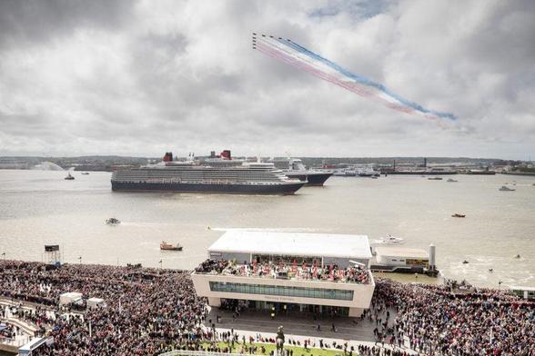 A large crowd gathers on the waterfront to watch a maritime event featuring multiple ships on the river, including the Queen Mary 2, while military jets fly overhead leaving trails of red, white, and blue smoke against a cloudy sky.