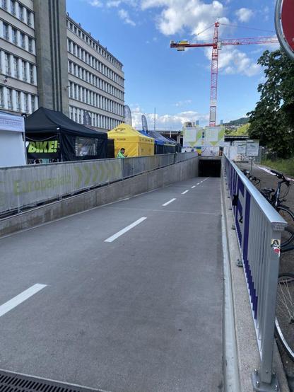 Une entrée de tunnel cyclable, à gauche les tentes d'une fête, en arrière fond une grue sur un ciel bleu