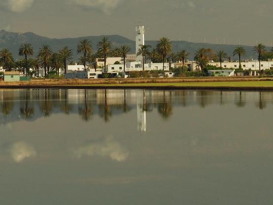 En la imagen vemos la localidad de Poblenou del Delta reflejada en un arrozal inundado de agua. Poblenou del Delta es una localidad perteneciente al municipio de Amposta, en la comarca  del Montsià de Tarragona. Vemos la torre de la iglesia, sus casitas blancas, sus palmeras, el cielo con pequeñas nubes y a lo lejos las montañas,  reflejadas en el agua. 