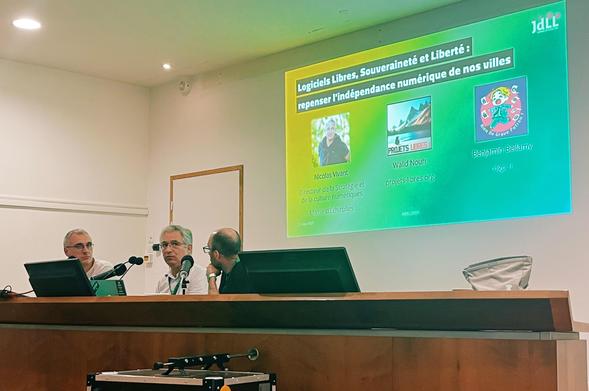 A photo of a stage in an auditorium with three men sitting behind a large podium and behind them there is a large screen with the title (in French): FOSS, Sovereignty and Freedom: rethinking the digital independence of our towns 