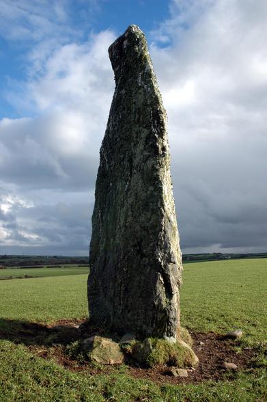 A tall standing stone of Bronze Age date in a wintry sunny sky. Lichen covers some of the stone, which stands in a grassy agricultural field.