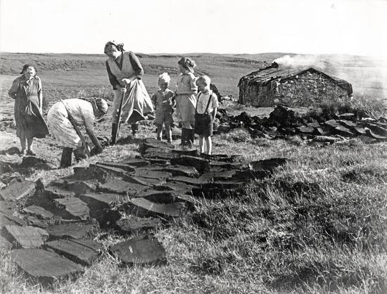Two women digging for peat, while an older woman and three children stand watching. Piles of cut peat lie in the foreground and middleground. In the background is a small thatched building with smoke pouring out that served as shelter during the summer months when peat-cutting for fuel was a primary occupation, readying for the winter.