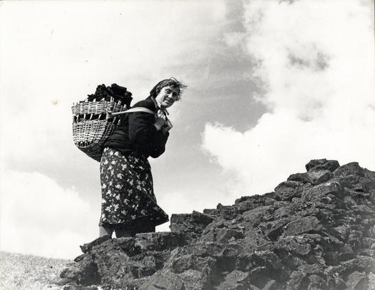 A woman, silhouetted against the sky, smiling for the camera, is climbing up a huge pile of cut peat, with a large basket full of peat on her back.