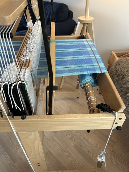 A side view of a full cloth beam on a table loom showing blue checked fabric ready to cut off the loom