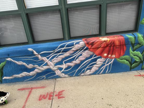 A colorful mural featuring a large red flower and flowing white tendrils on a blue background, alongside green leaves. The bottom part of the image shows a sidewalk with a partially visible shoe on roller skates and red graffiti text.