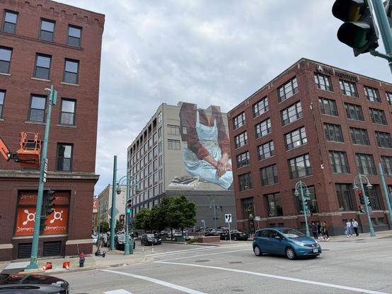 Urban scene featuring a large mural on a building depicting a person in an apron working with hands. Nearby structures include red brick buildings and a traffic signal. The area appears to be bustling with vehicles and pedestrians.