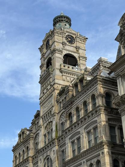 The image shows a historic building featuring a prominent clock tower with a green dome. The architecture includes decorative elements and arched windows, with the inscription "Chamber of Commerce" visible on the facade. The sky is clear with some clouds.