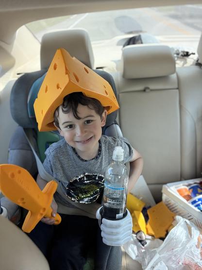 A child sits in a car seat wearing a large piece of orange cheese-shaped headwear. They hold a foam sword in one hand and a water bottle in the other, while smiling. The car interior shows some clutter in the background.