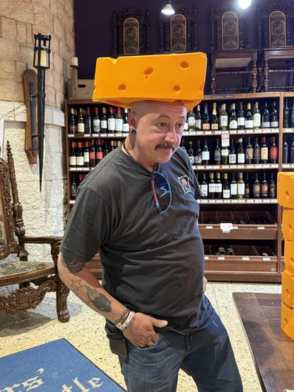 A man with tattoos wears a large, bright yellow piece of cheese on his head, standing in a store filled with bottles of wine. The setting includes wooden shelves and ornate chairs in the background.