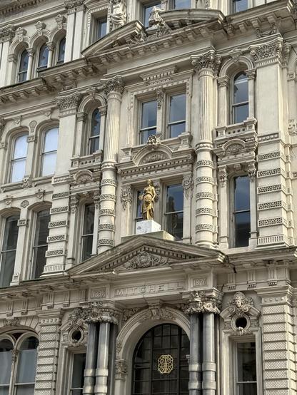An ornate building facade featuring intricate architectural details, including several decorative columns and sculptures. A statue atop the building is visible, with the name "MITCHELL" inscribed above the entrance, along with the years "1839" and "1876