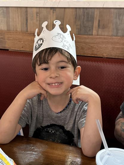 A young child with brown hair wearing a paper crown sits at a table, smiling with hands resting on their cheeks. The child is dressed in a gray t-shirt and has a beaded necklace. A drink with a straw is visible on the table,